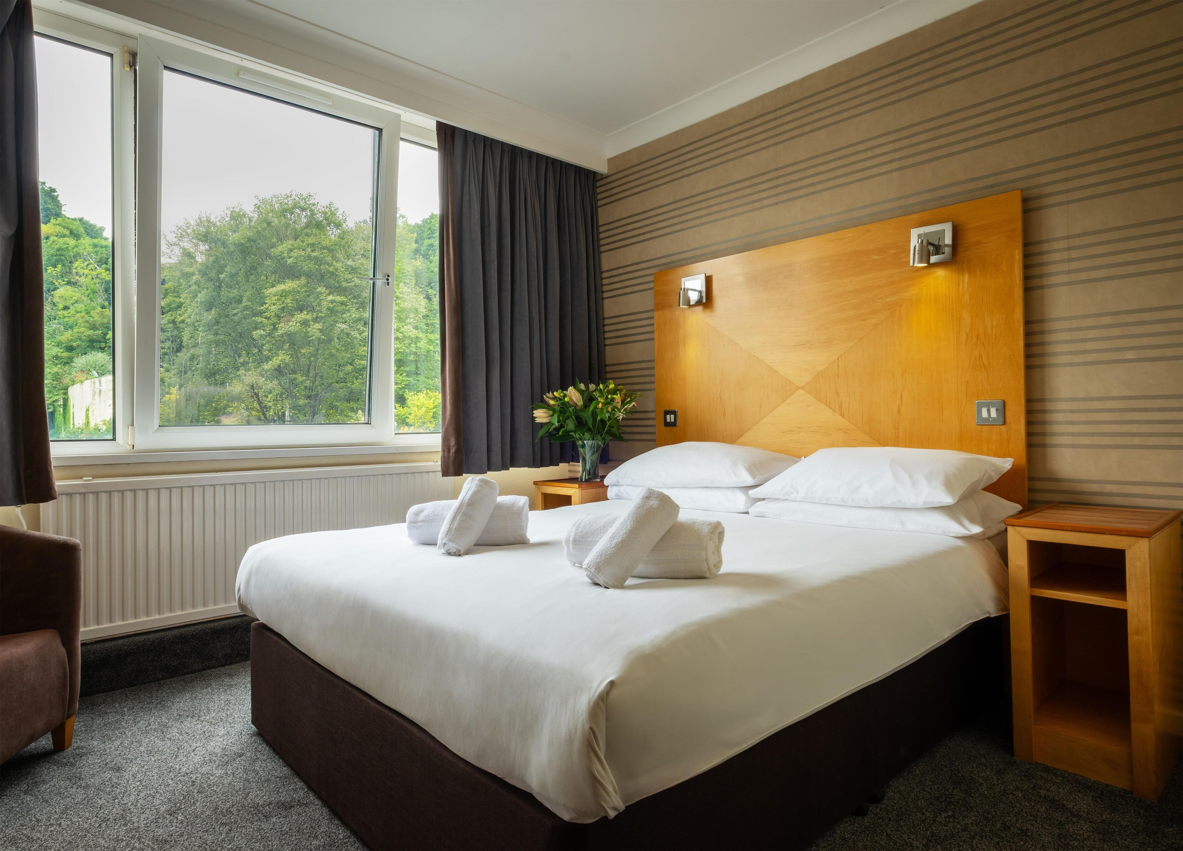 Habitación de hotel con una cama grande, una mesita de noche, cortinas y vistas a árboles.