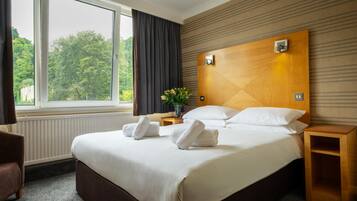 Habitación de hotel con una cama grande, una mesita de noche, cortinas y vistas a árboles.