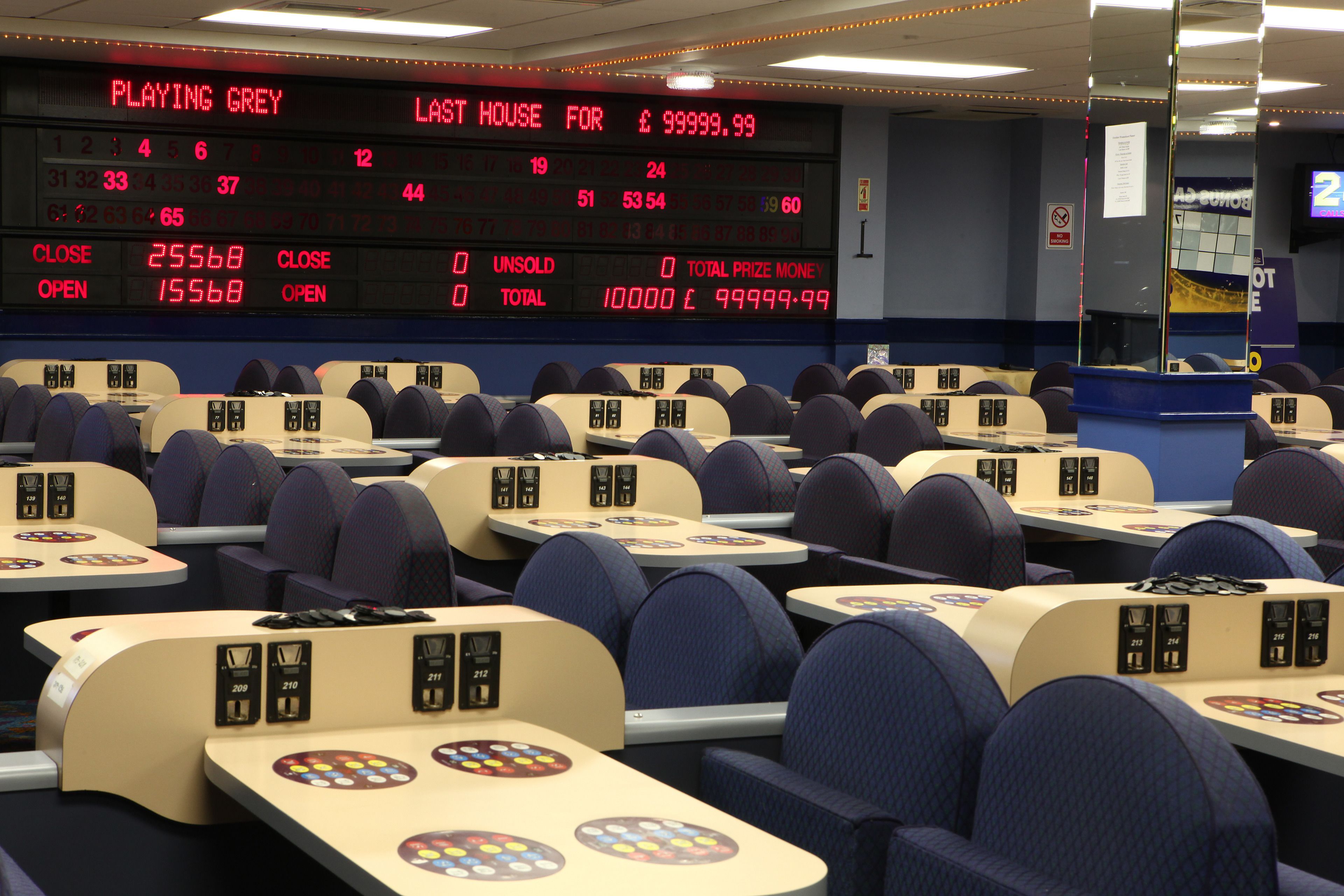 Un casino con múltiples mesas de juego y sillas, un marcador digital que muestra los resultados y una zona de estar azul.