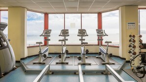 Un gimnasio con cintas de correr y pesas, ventanas amplias con vista al mar y techo con ventilación.