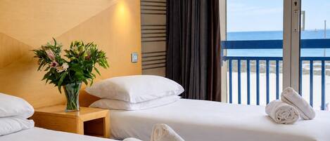 Une chambre d’hôtel avec deux lits, une table de chevet ornée d’un vase de fleurs et une vue sur la mer depuis un balcon.