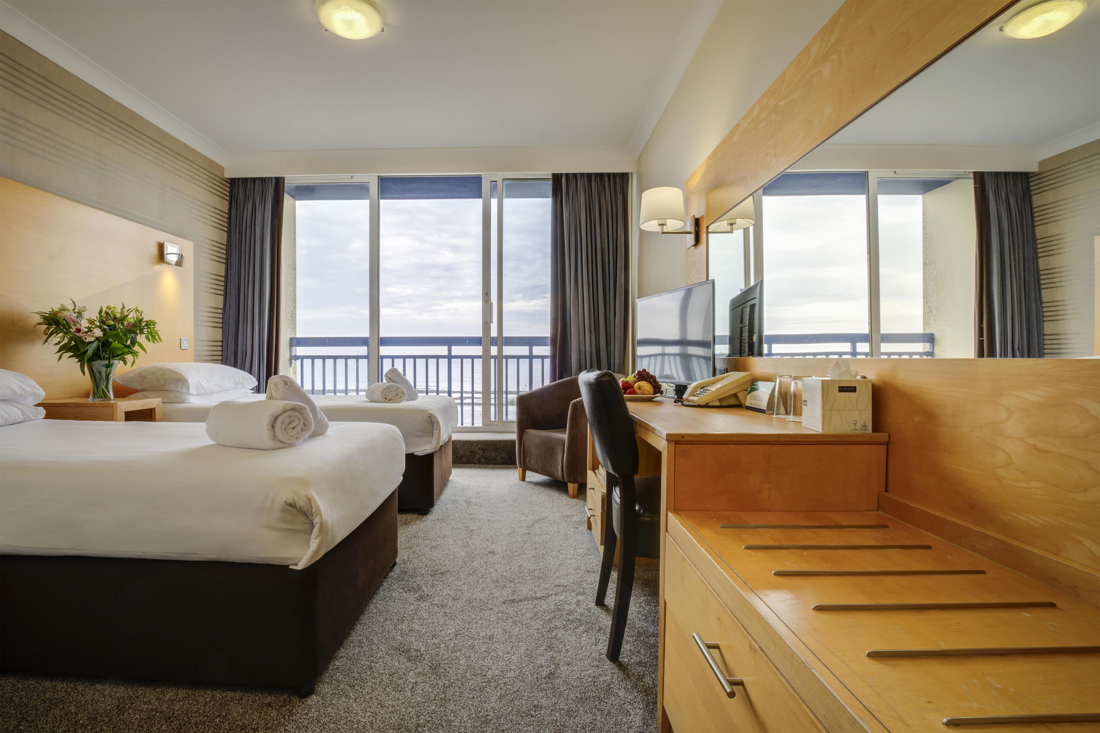 A hotel room with a bed, a desk, a chair, a TV, and a view of the sea.