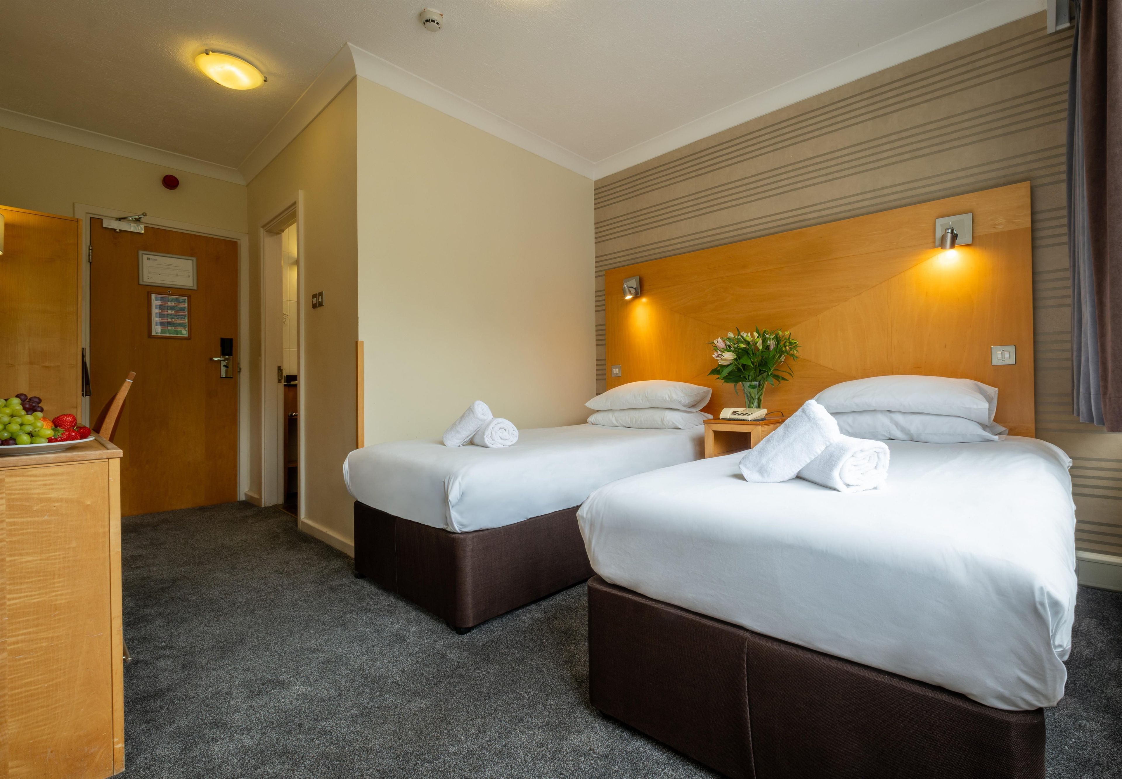 A hotel room with two beds, a desk with fruit, a plant, and a wooden headboard.