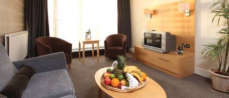 A hotel room with a sofa, two armchairs, a small table with fruit, a TV, and a potted plant.