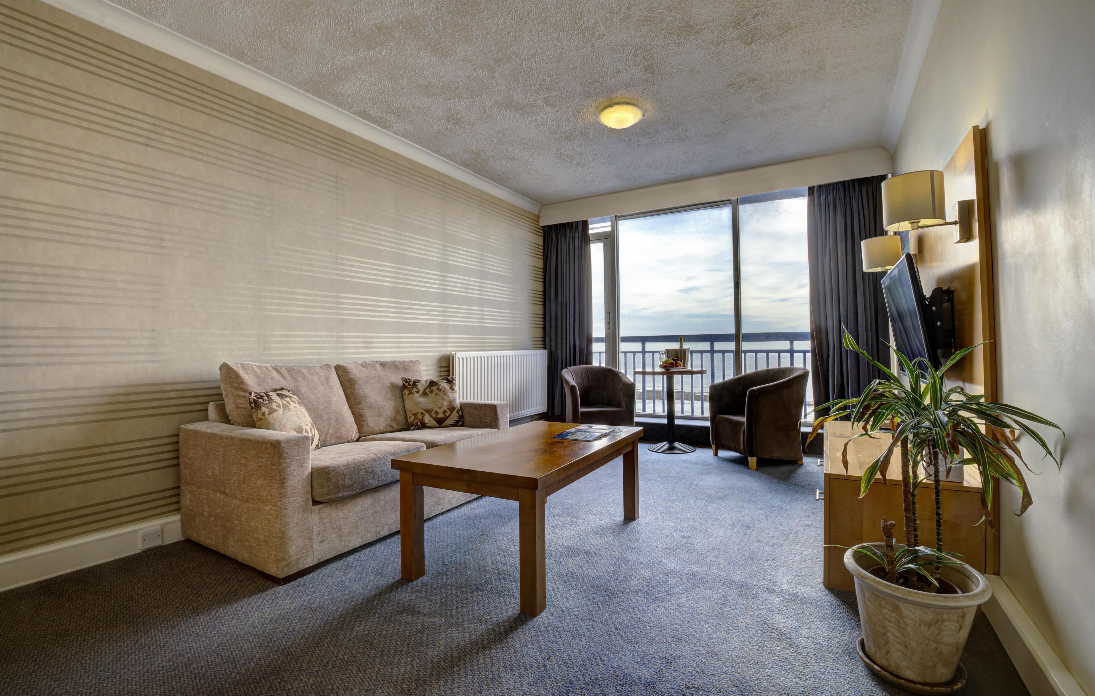 Habitación de hotel con sofá, una mesa de madera, planta en maceta, balcón con vistas y un ventanal con cortinas.