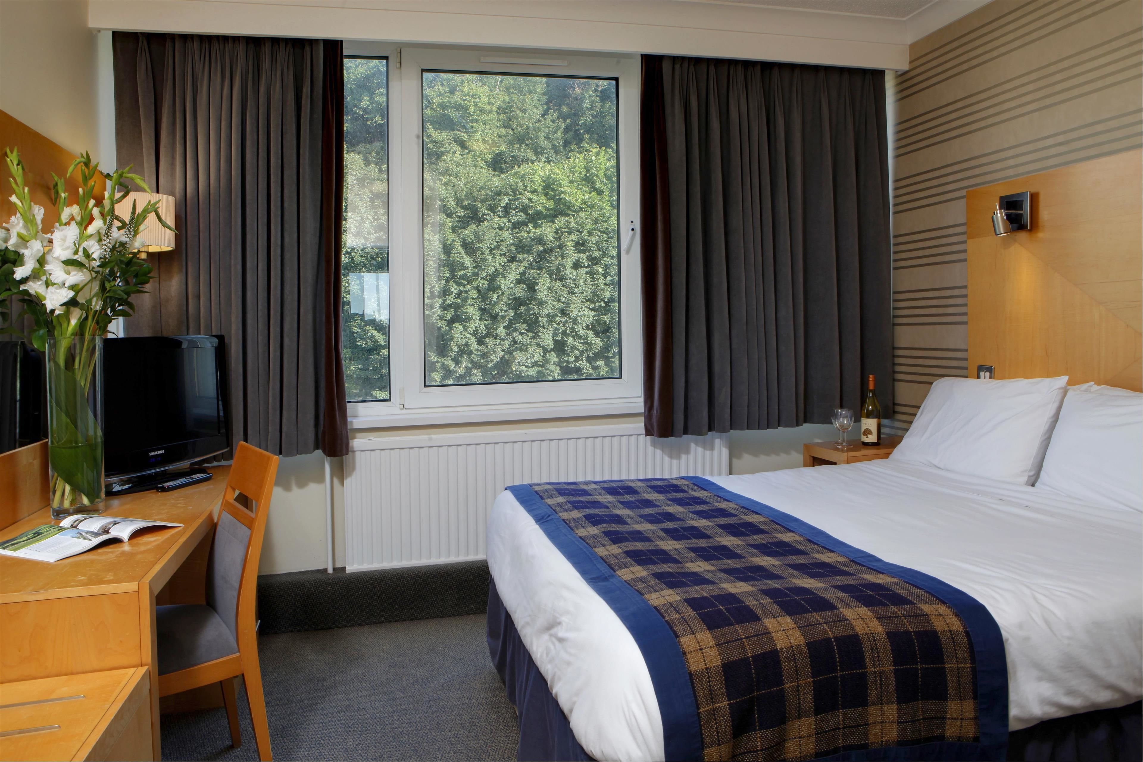 Habitación de hotel con cama, escritorio y silla, televisor, ventana con cortinas y vistas a árboles.