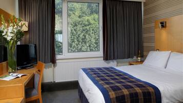 Habitación de hotel con cama, escritorio y silla, televisor, ventana con cortinas y vistas a árboles.
