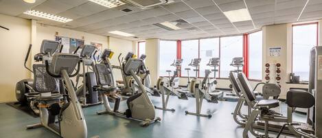 A gym with various exercise machines, large windows, and a clean floor.