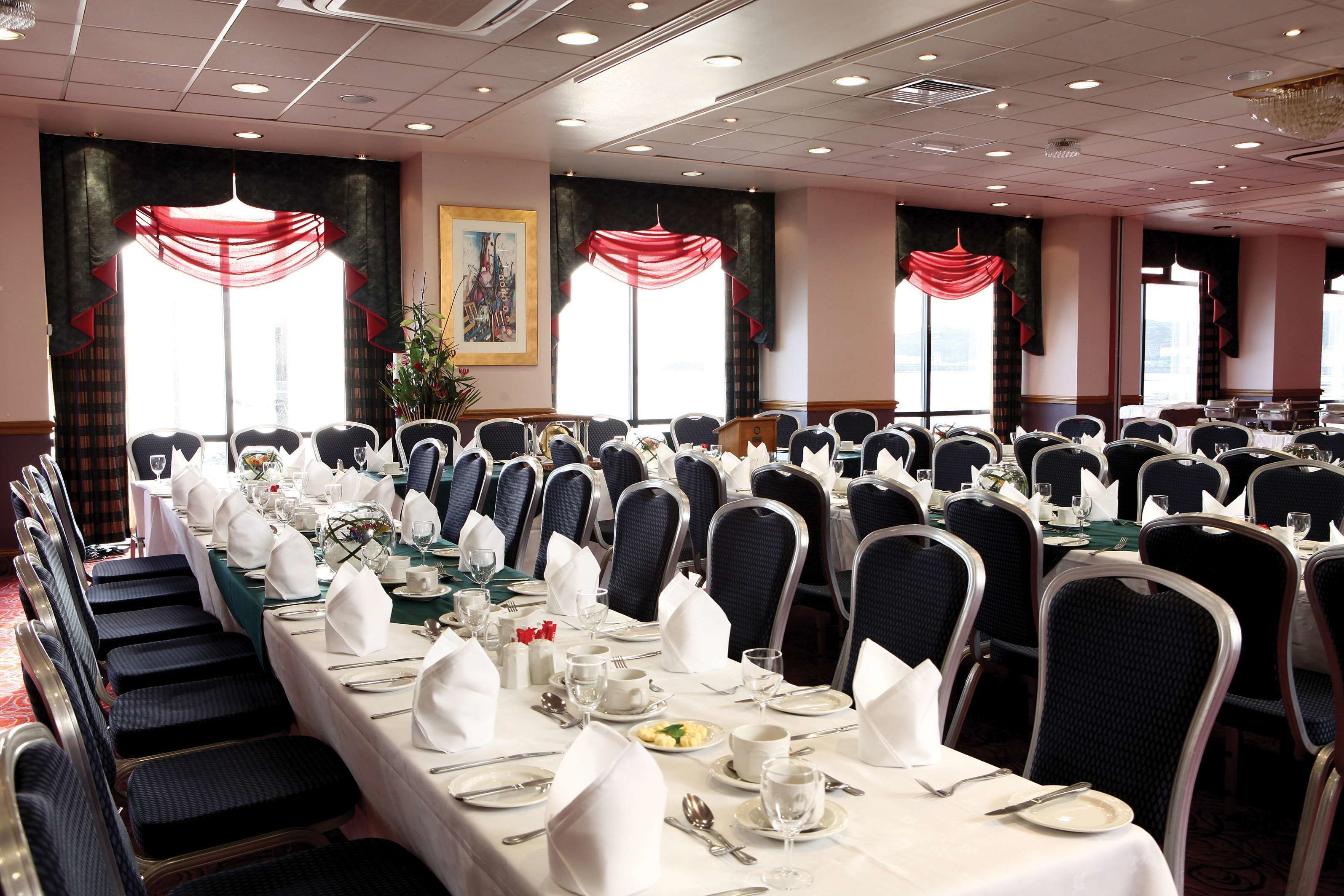 Une salle de banquet où de longues rangées de tables sont dressées pour le repas, chacune étant recouverte d’une nappe blanche et agrémentée de chaises et de couverts soigneusement disposés.