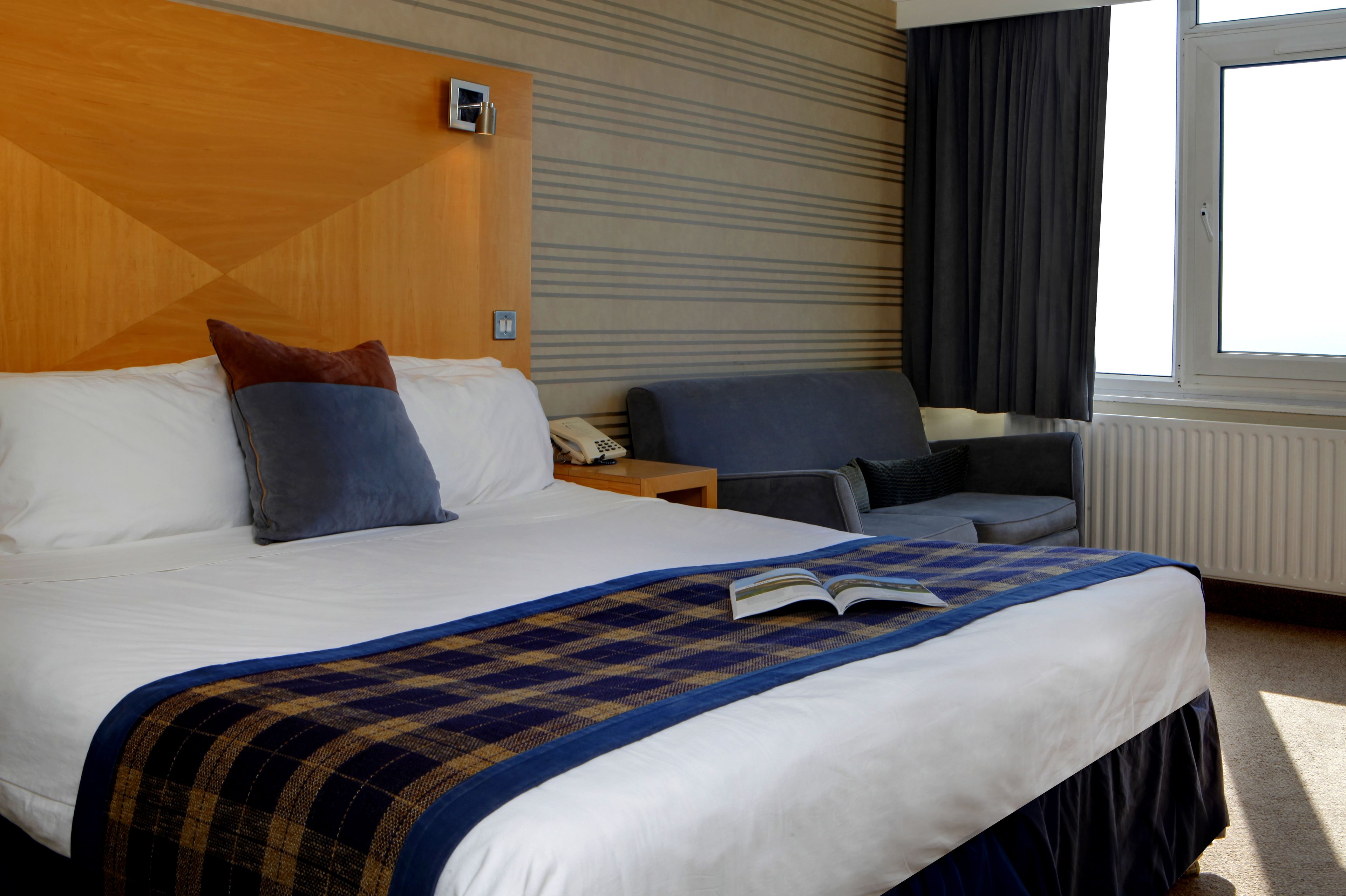 A hotel room with a large bed, a bedside table with a phone, a chair, and a window with curtains.