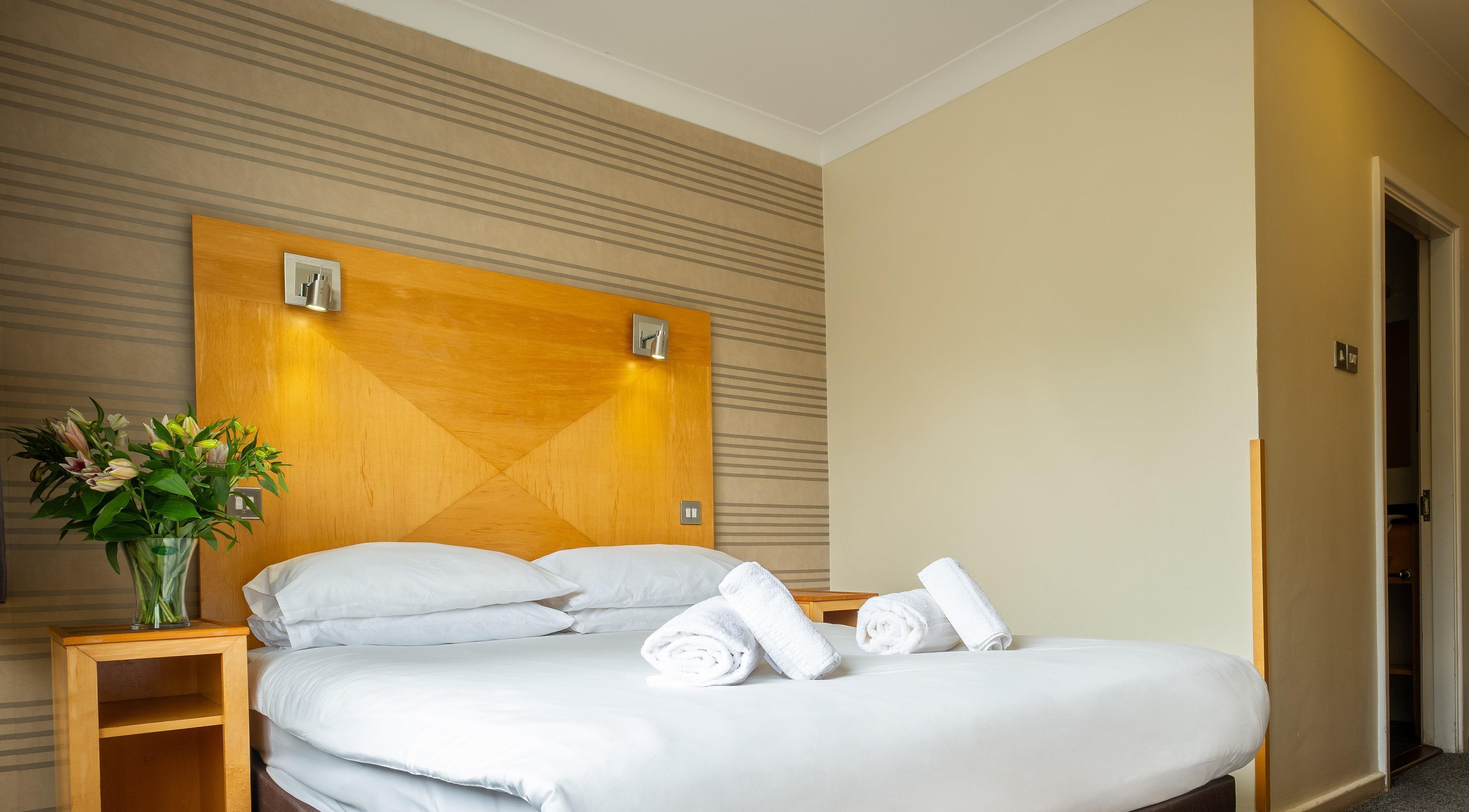 Una habitación de hotel con una cama, una mesita de noche, un florero con flores y un cabecero de madera.