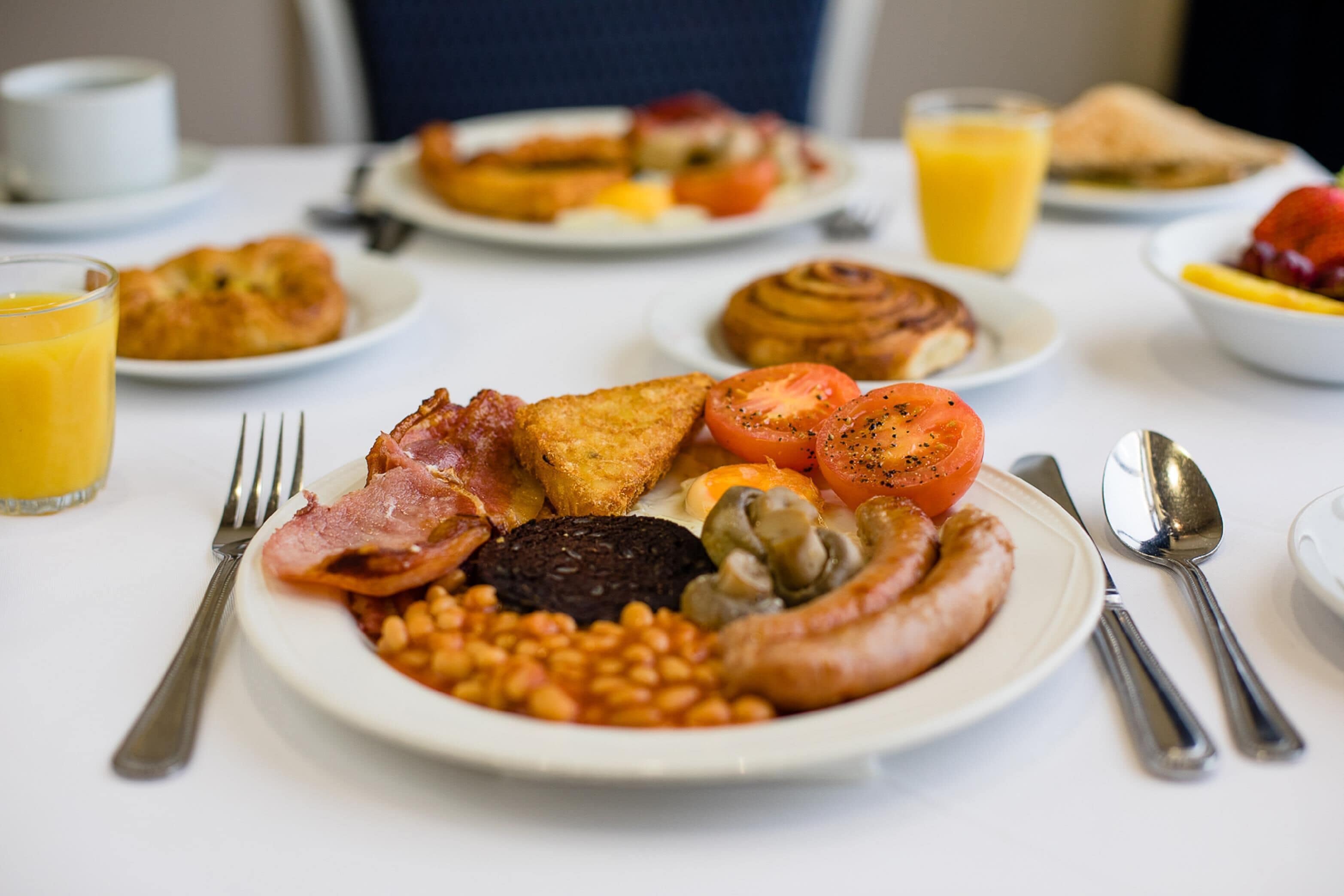 Ein Frühstückstisch mit Tellern voller Speisen, darunter Eier, Speck, Würstchen, Bohnen und Toastbrot.