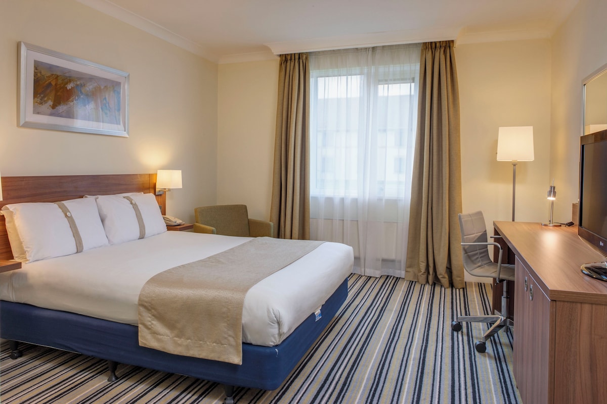 Habitación de hotel con una cama grande, un escritorio, una silla, un televisor y un ventanal con cortinas.