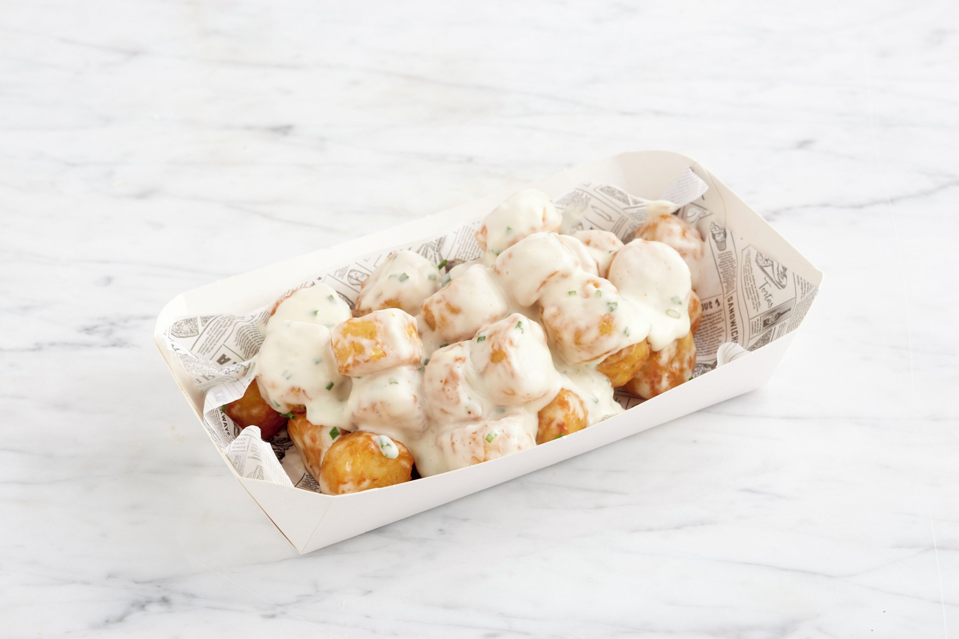 A tray of food items, possibly fritters, covered in a creamy sauce, placed on a white surface with a newspaper-like liner.