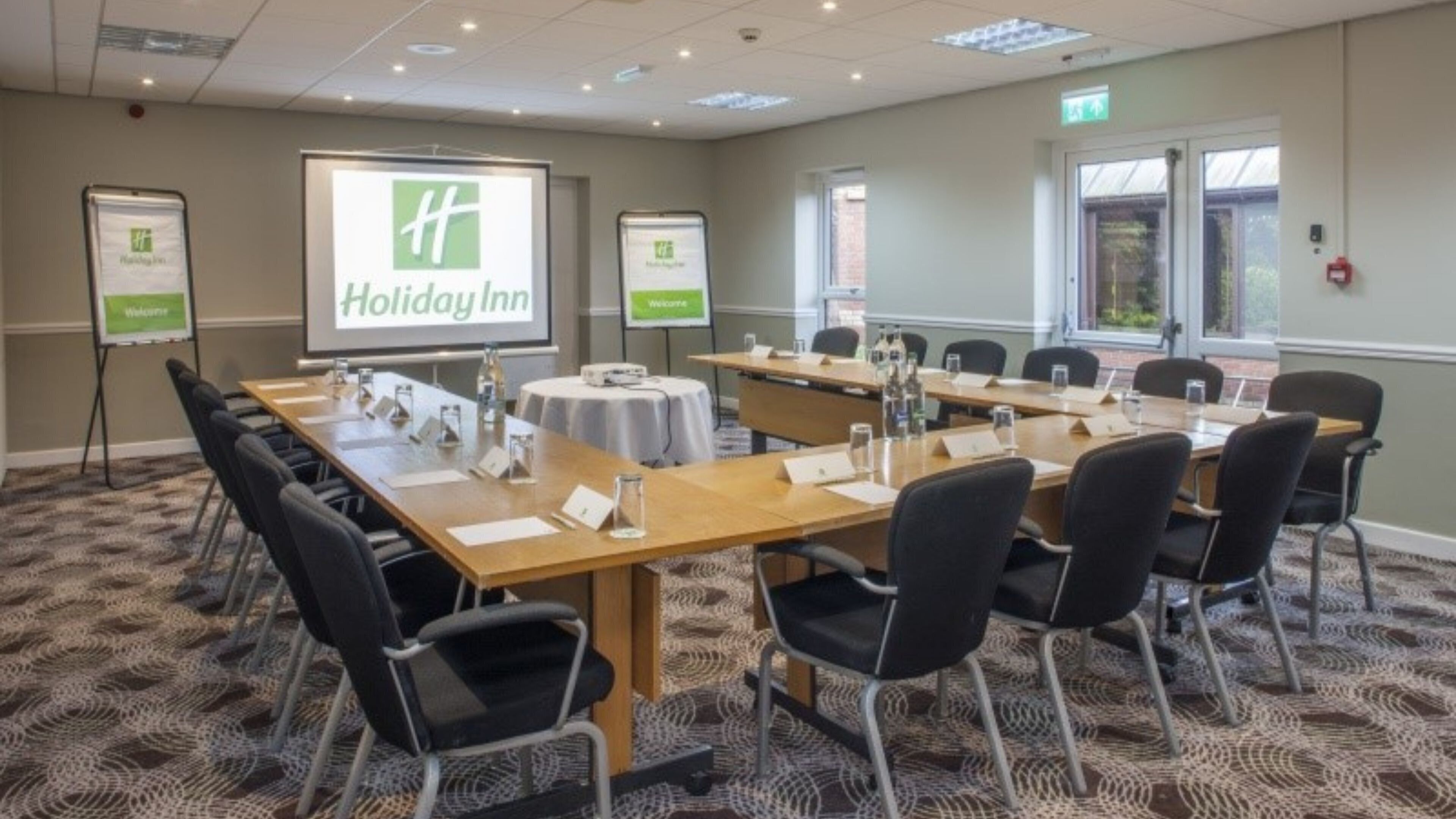 Et mødelokale med et langt bord, stole, navneskilte og et projektorskærm der viser Holiday Inn logoet.
