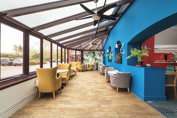 Un restaurante con una larga mesa de madera y sillas amarillas, una pared azul con mostrador rojo y techo de vidrio.