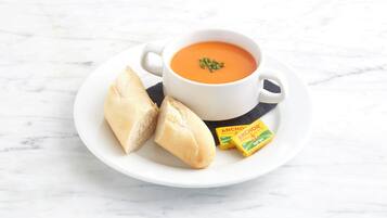 Una taza de cerámica blanca con sopa y un asa, acompañada de dos rebanadas de pan y dos paquetes de mantequilla sobre un plato blanco.