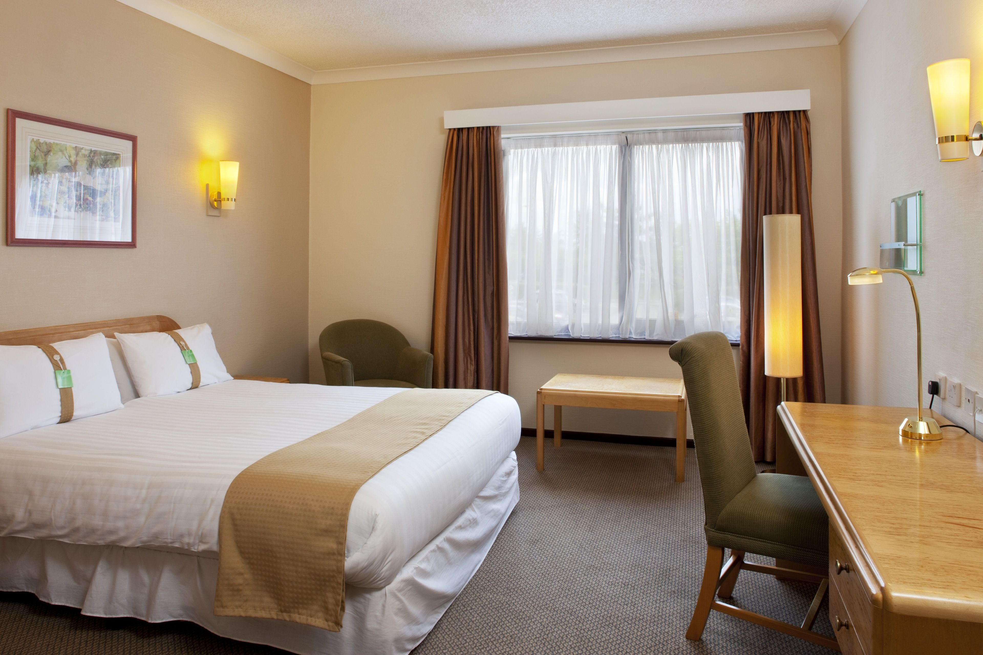 A hotel room with a large bed, a desk, a chair, a lamp, and a window with curtains.