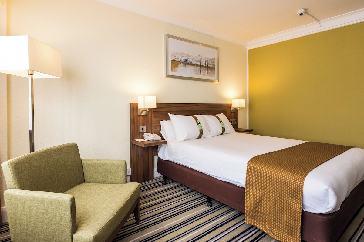 Una habitación de hotel con una cama, una silla, una mesita de noche y un cuadro en la pared.