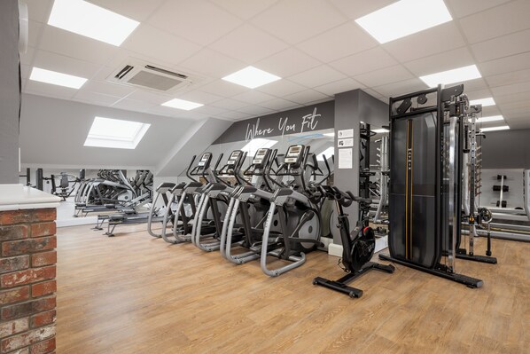 Une salle de sport équipée de nombreuses machines d’exercice, avec un mur en briques et un plafond agrémenté de lucarnes.