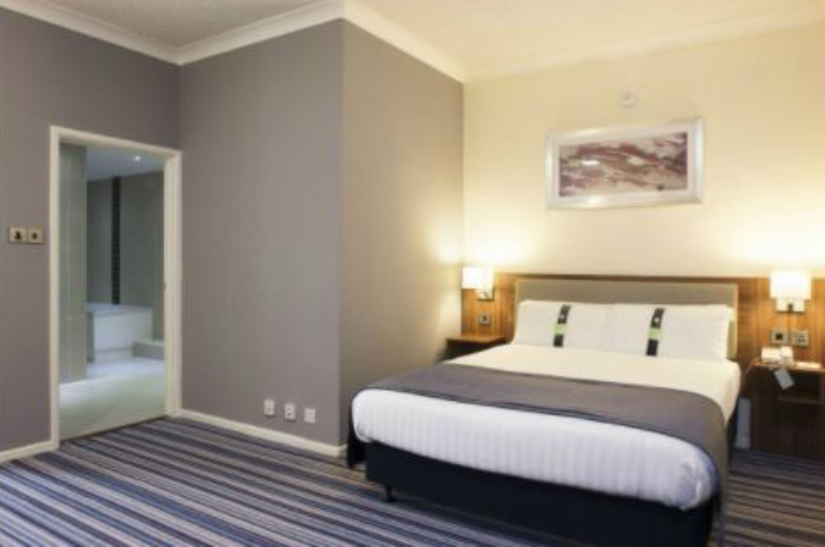 Una habitación de hotel con una cama grande, una mesita de noche, una lámpara de pared y una puerta que da a un baño.
