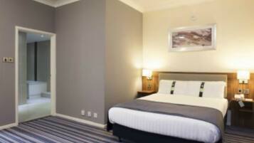 Una habitación de hotel con una cama grande, una mesita de noche, una lámpara de pared y una puerta que da a un baño.