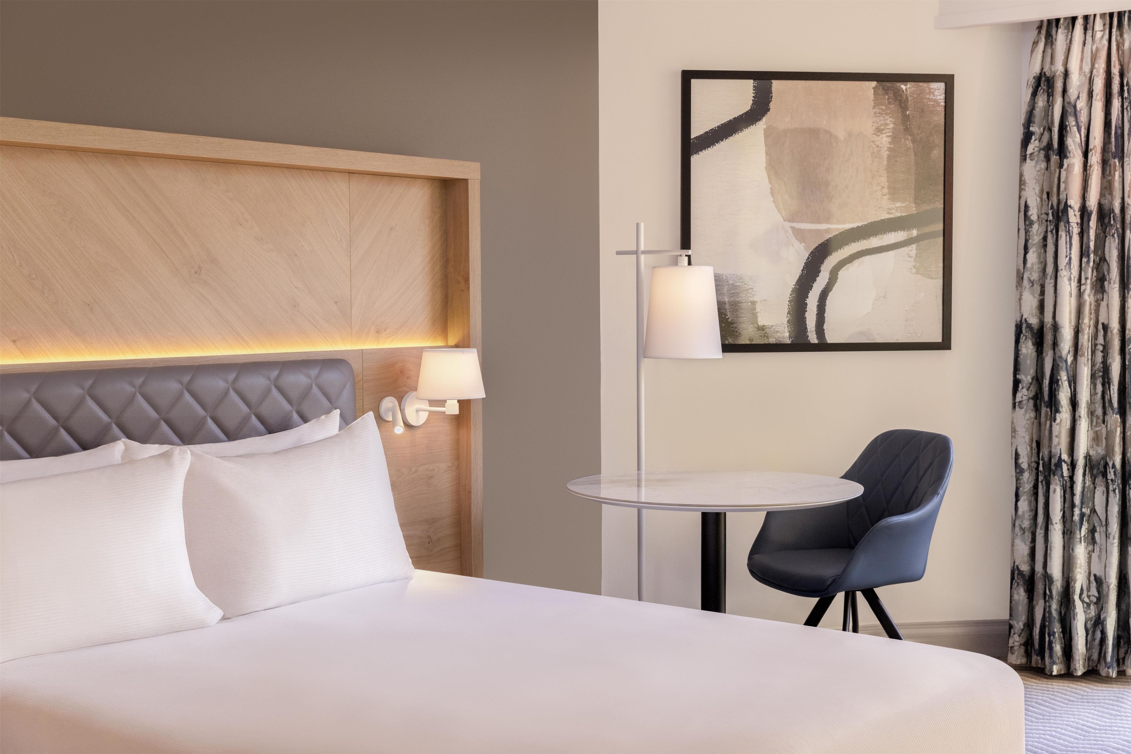 A hotel room with a large bed, a bedside table with a lamp, a chair, and a painting on the wall.