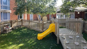 A wooden play structure with a slide, surrounded by a brick building and a tree.