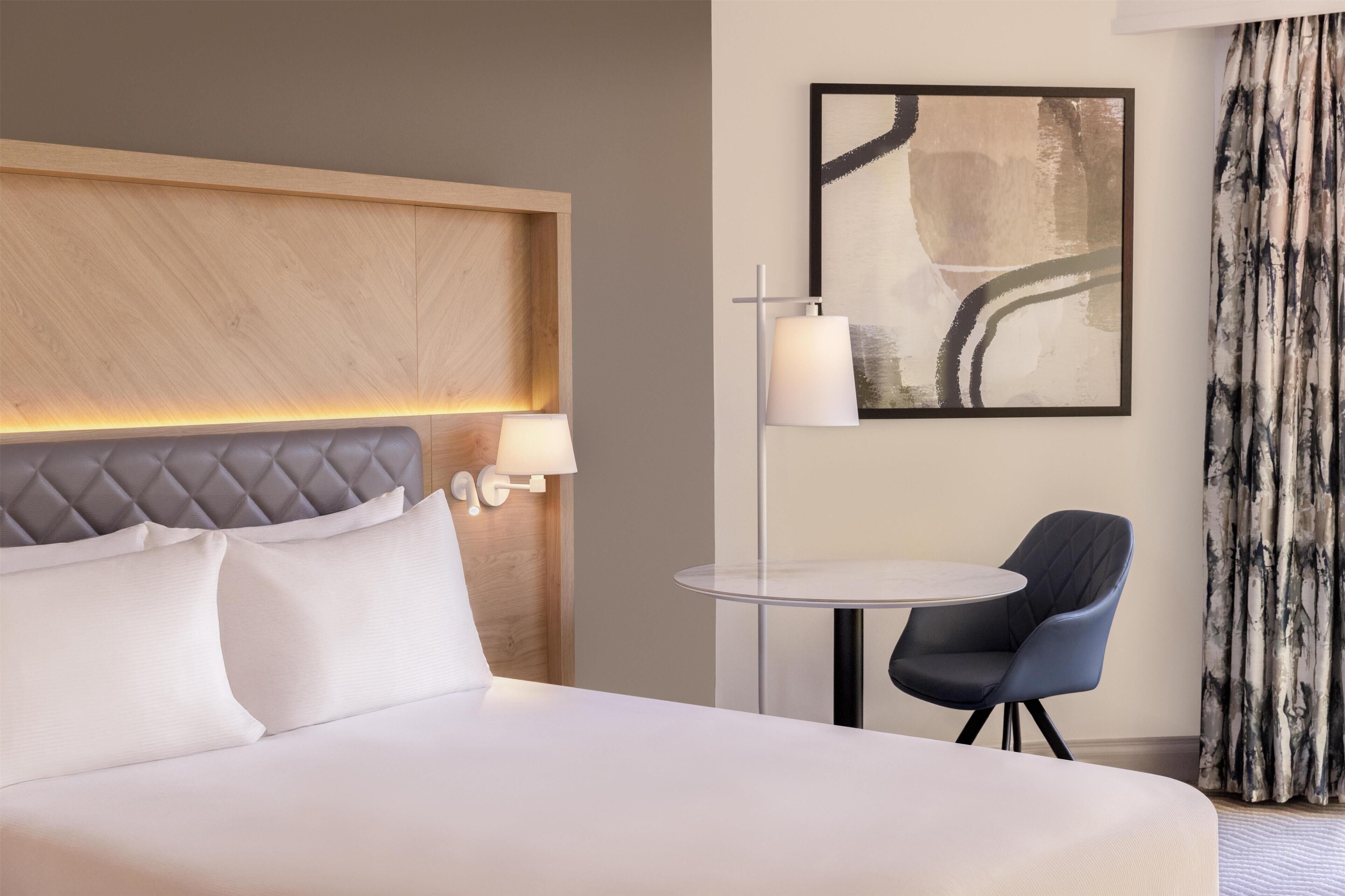 A hotel room with a bed, a round table, a chair, a wall-mounted lamp, and a framed abstract painting.