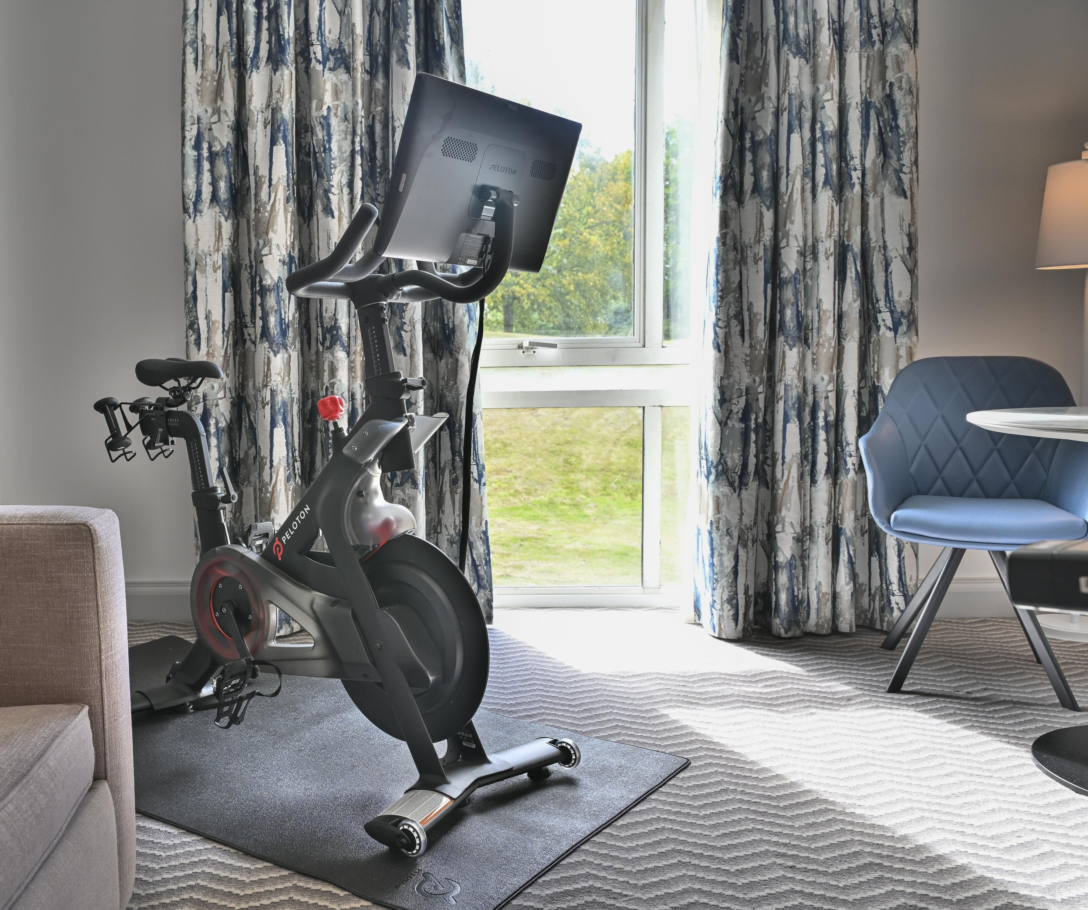 Una bicicleta estática Peloton en una habitación con un ventanal grande, un sillón azul y una alfombra estampada.