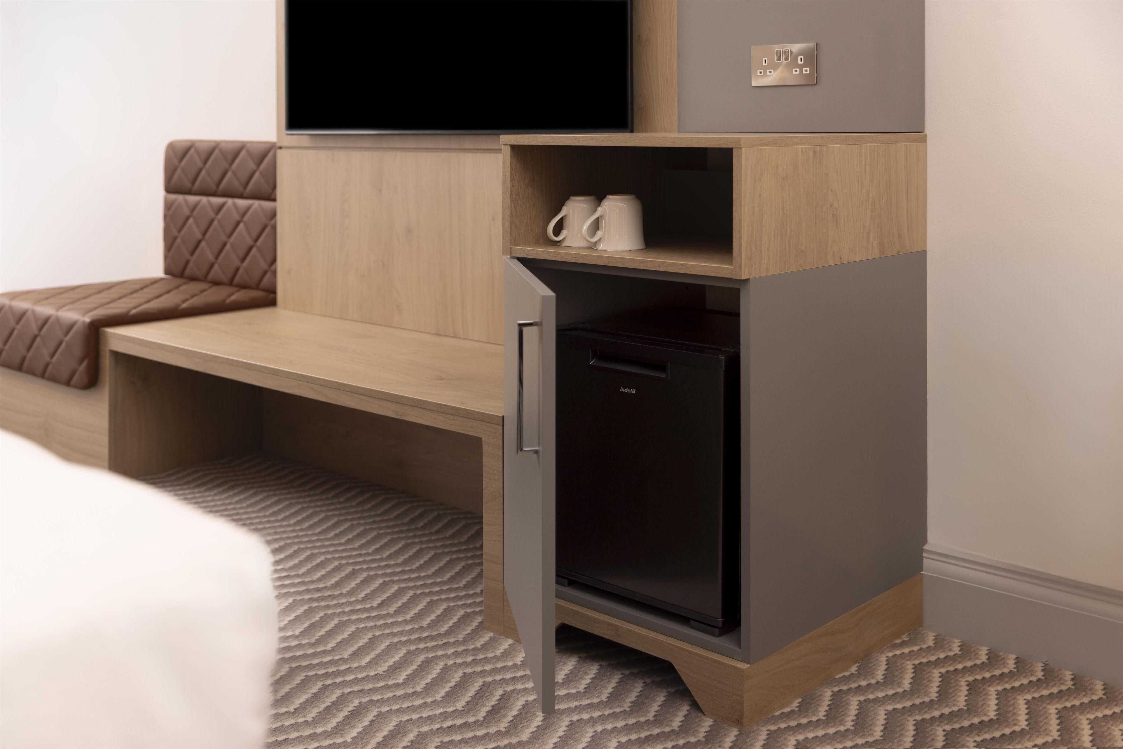 A compact room with a bed, a desk, a built-in TV, a mini-fridge, and a power outlet.