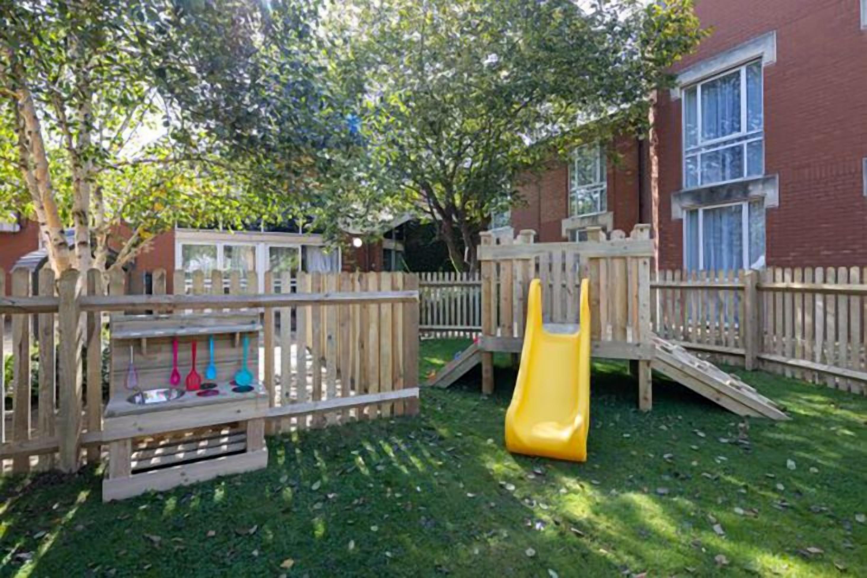 A playground with a slide and climbing structures, surrounded by a wooden fence, with a building in the background.