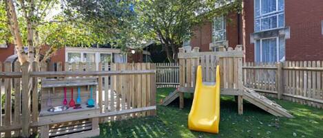 A playground with a slide and climbing structures, surrounded by a wooden fence, with a building in the background.