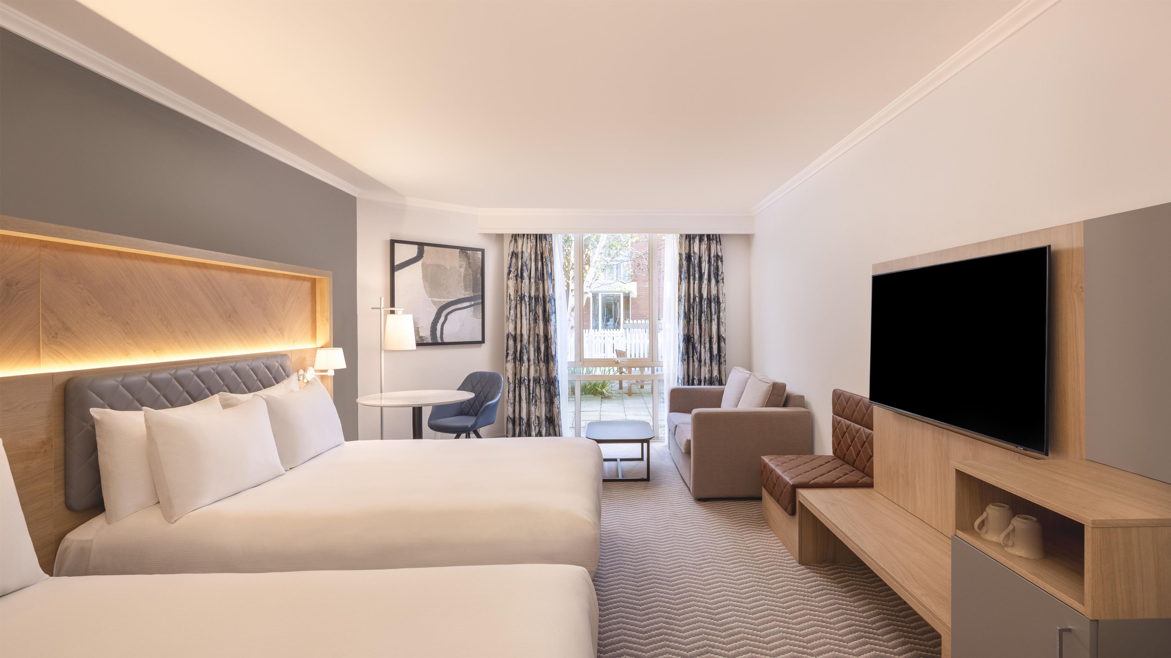 A hotel room with two beds, a TV, a desk, and a view of the outdoors.