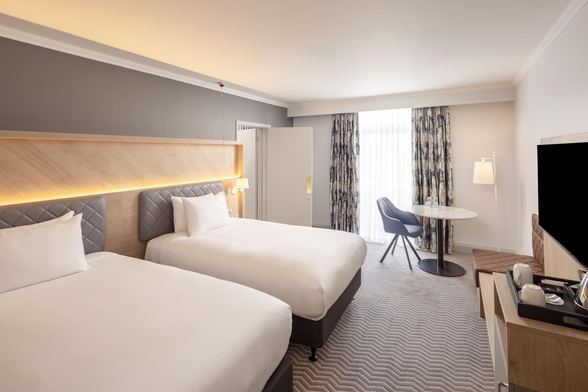 Quarto de hotel com duas camas, uma escrivaninha com cadeira, televisão e janela com cortinas.