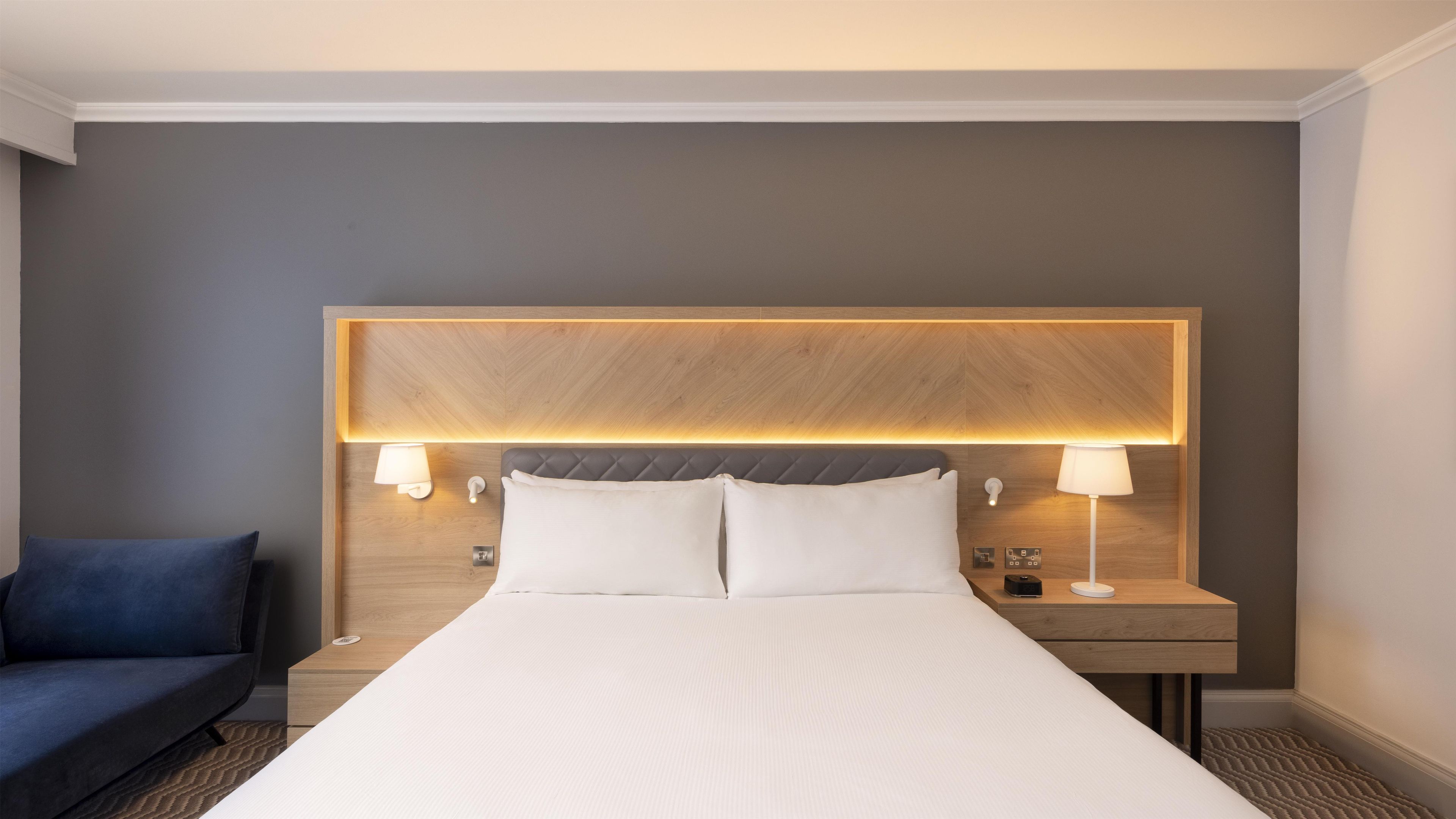 Habitació d'hotel amb un llit gran, dues làmpades de nit, un panell de fusta al capçal i un butacó blau.