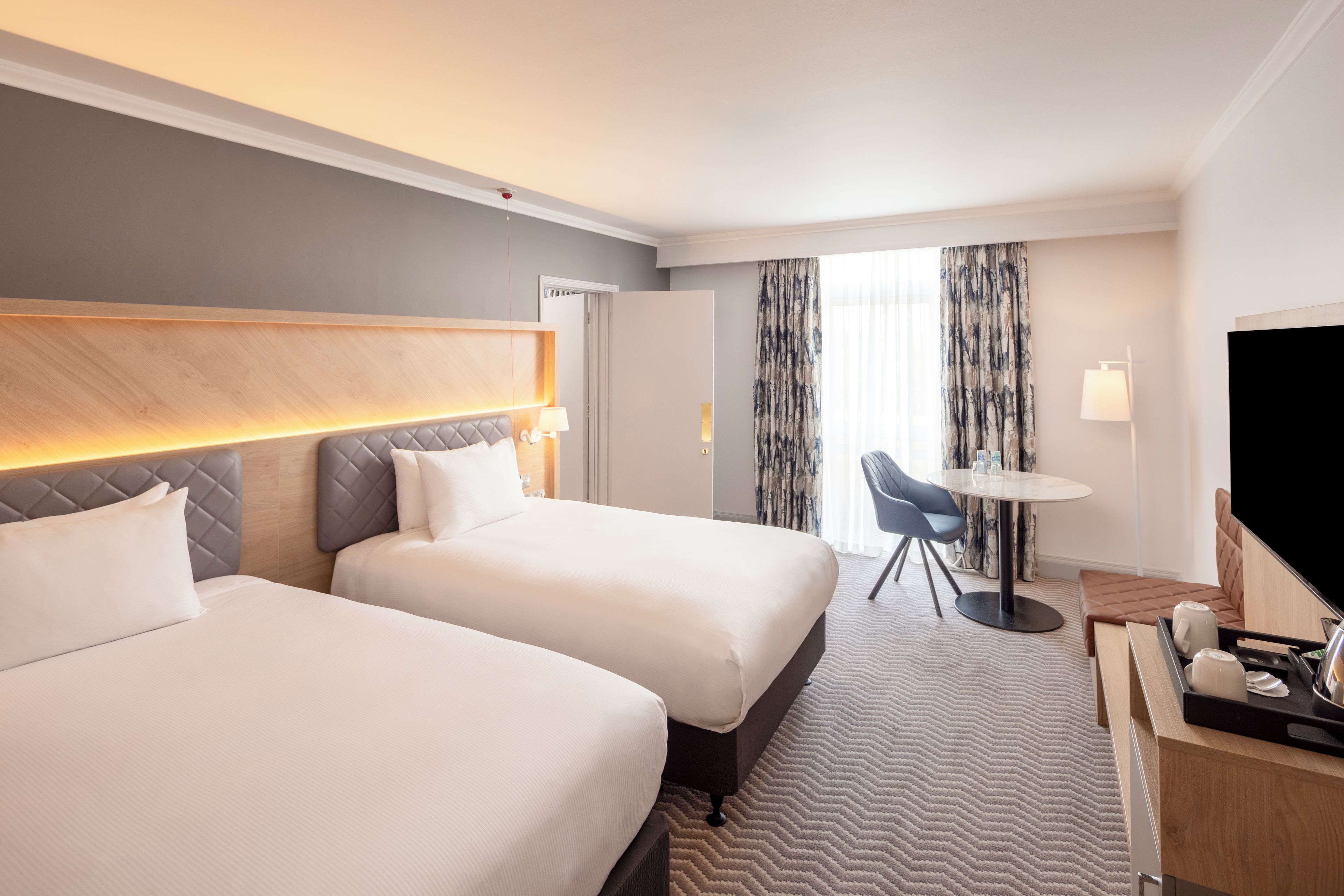 Habitación de hotel con dos camas, un escritorio con silla, televisor y ventana con cortinas.