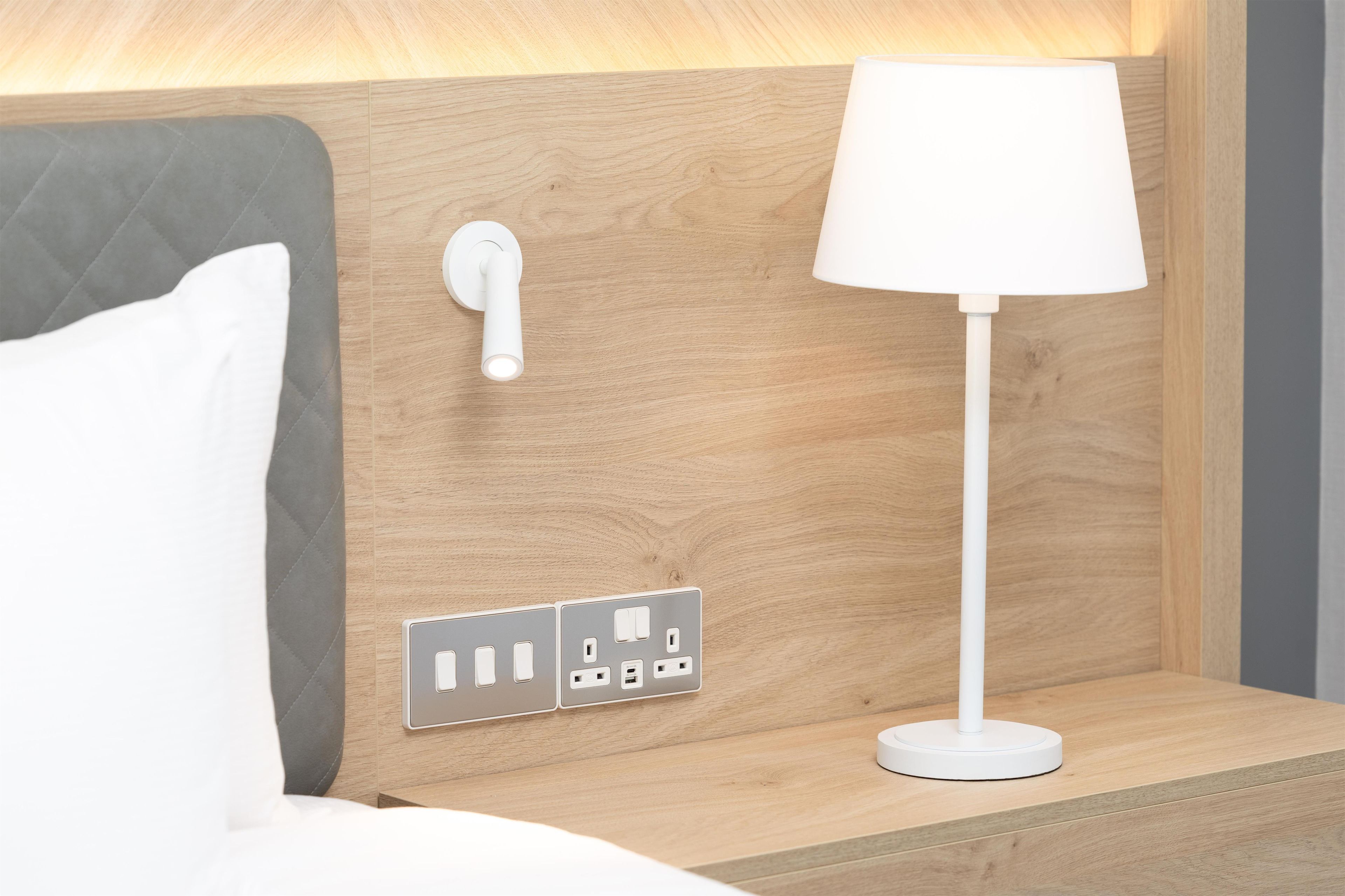A neatly made bed with a grey headboard, a white bedside lamp, and a wall-mounted power outlet with multiple sockets.
