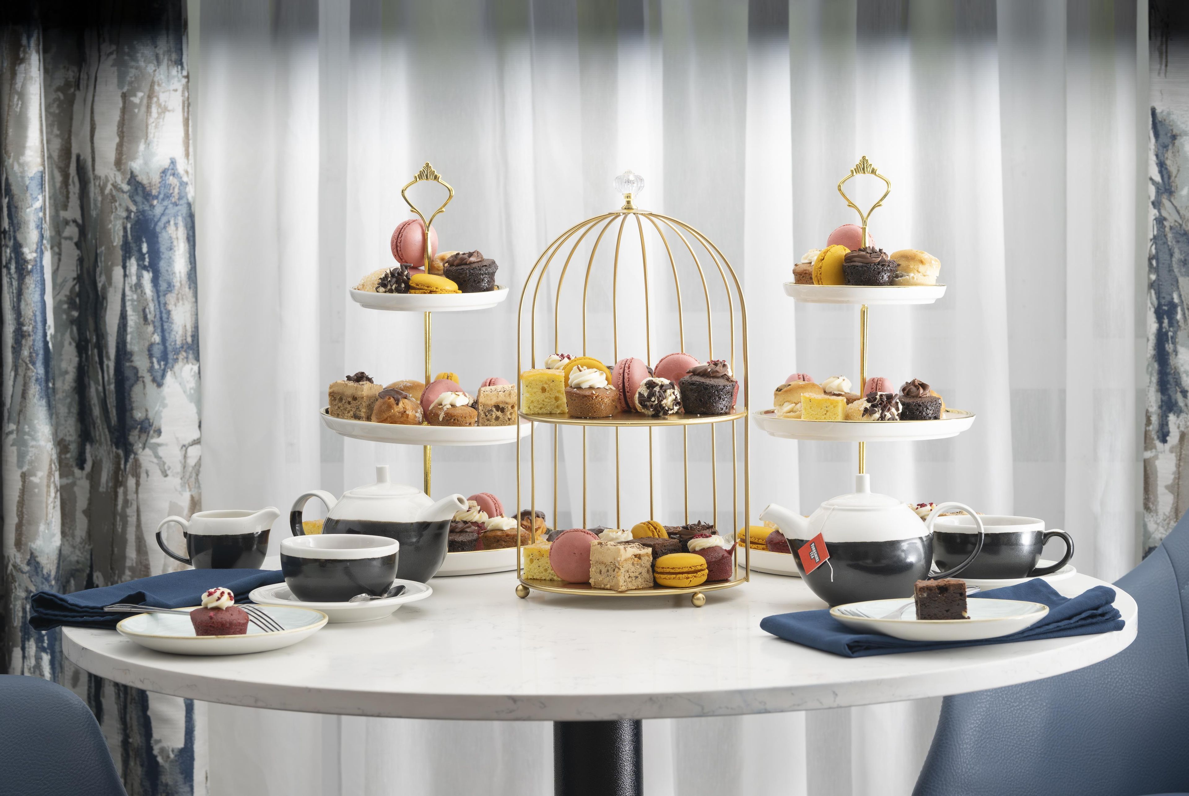 A three-tiered dessert stand with various pastries and macarons, accompanied by teapots and cups on a round table.