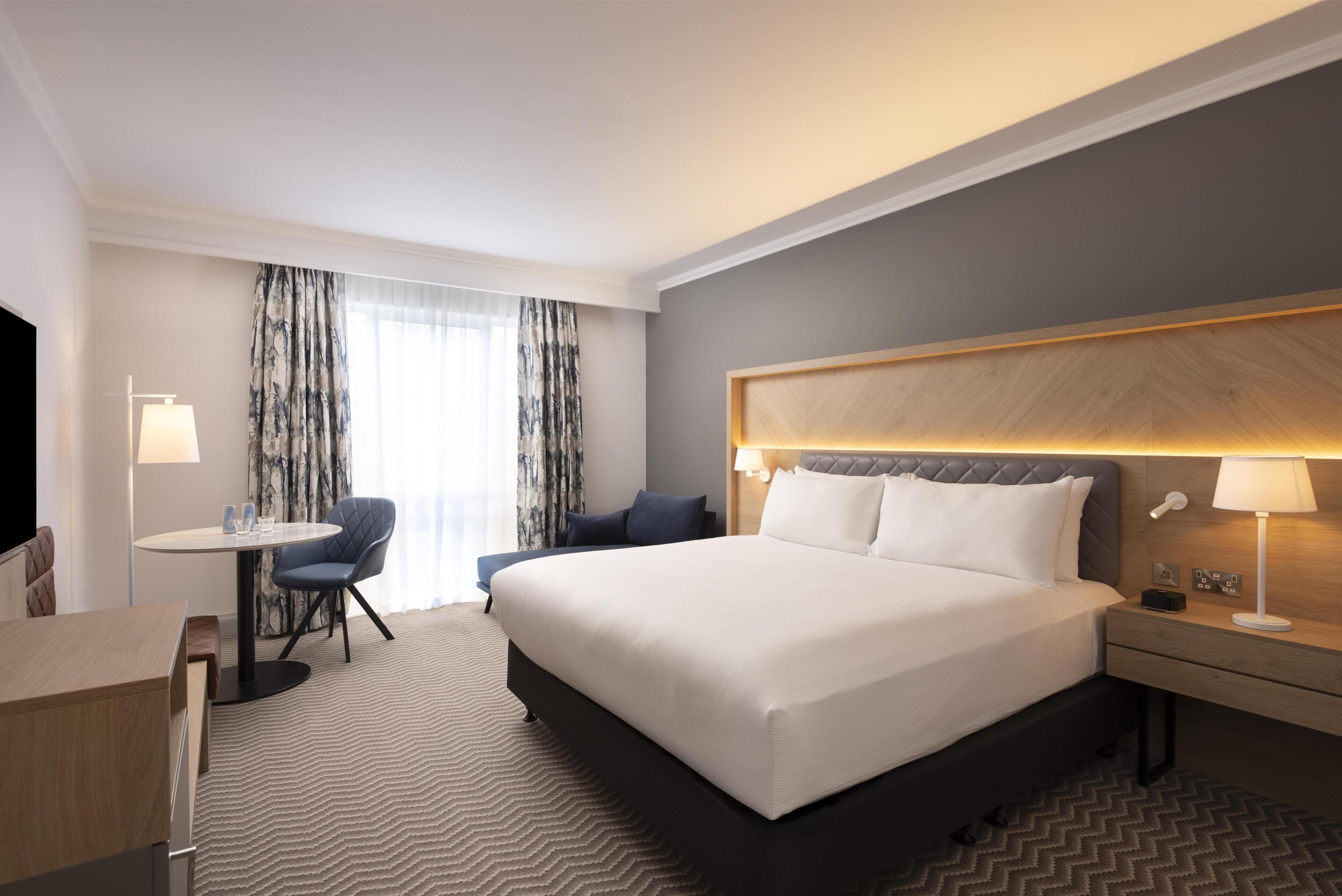 A hotel room with a large bed, a desk, a chair, a TV, and a window with curtains.