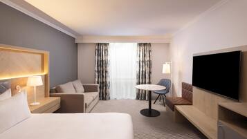 A hotel room with a bed, a desk with a lamp, a TV, a sofa, a chair, and a table.