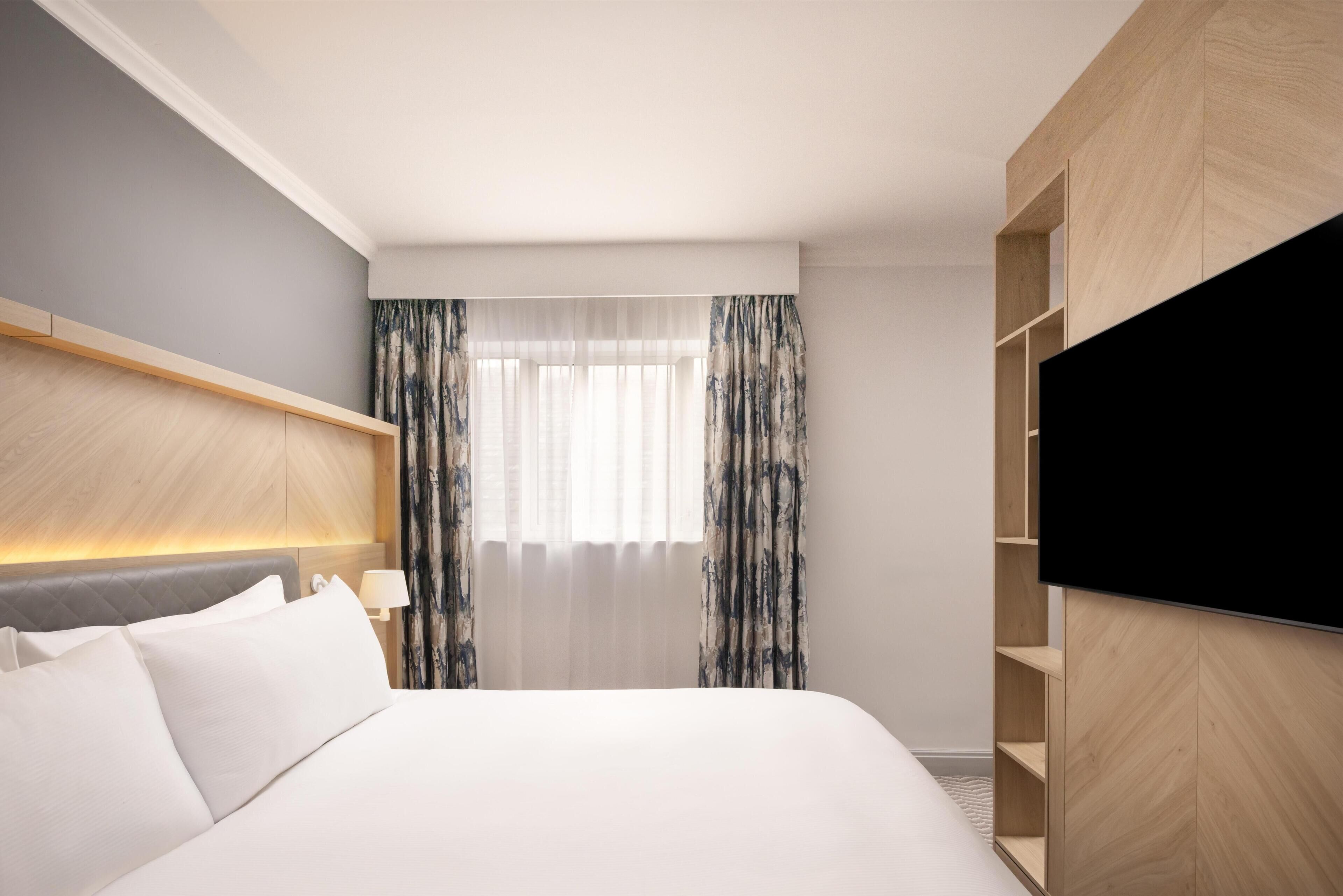 Habitación de hotel con cama, televisor, cabecero, ventana con cortinas y estanterías empotradas.