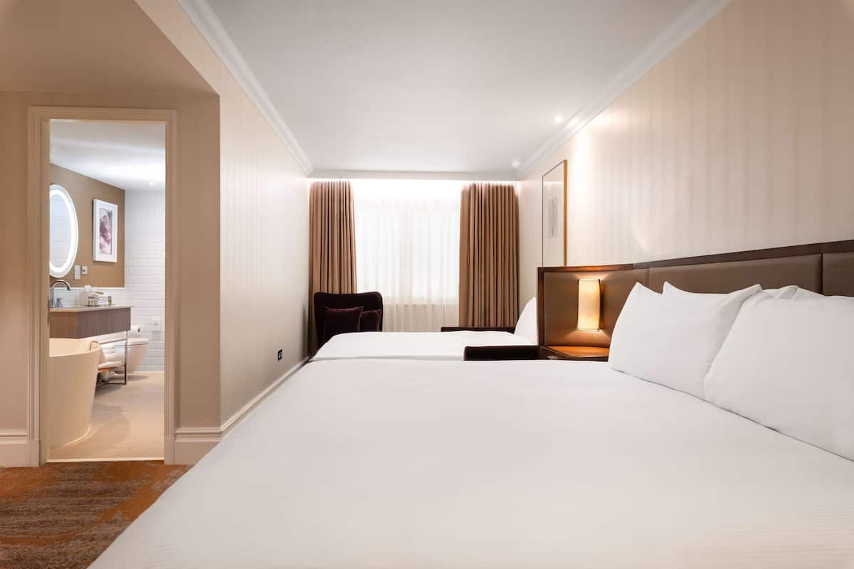 Una camera d'albergo con un letto grande, un comodino con lampada, una sedia, una scrivania e un bagno visibile tramite una porta aperta.