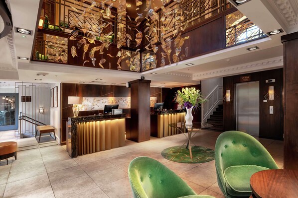 Um hall de hotel com balcão de recepção, cadeiras verdes, escada e teto decorativo.