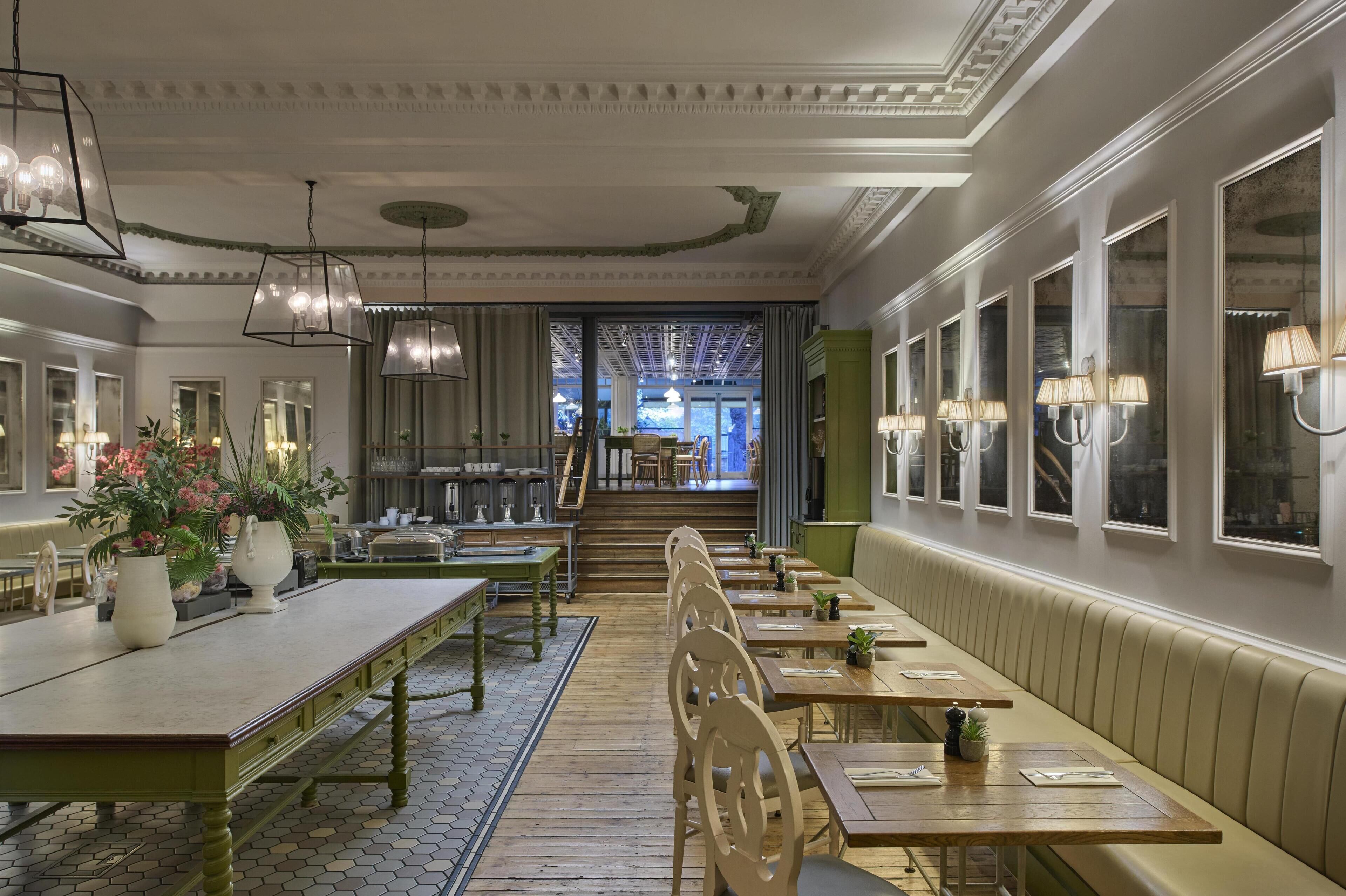 A restaurant with a long wooden bench seating area, a central staircase, and a view of the street through large windows.