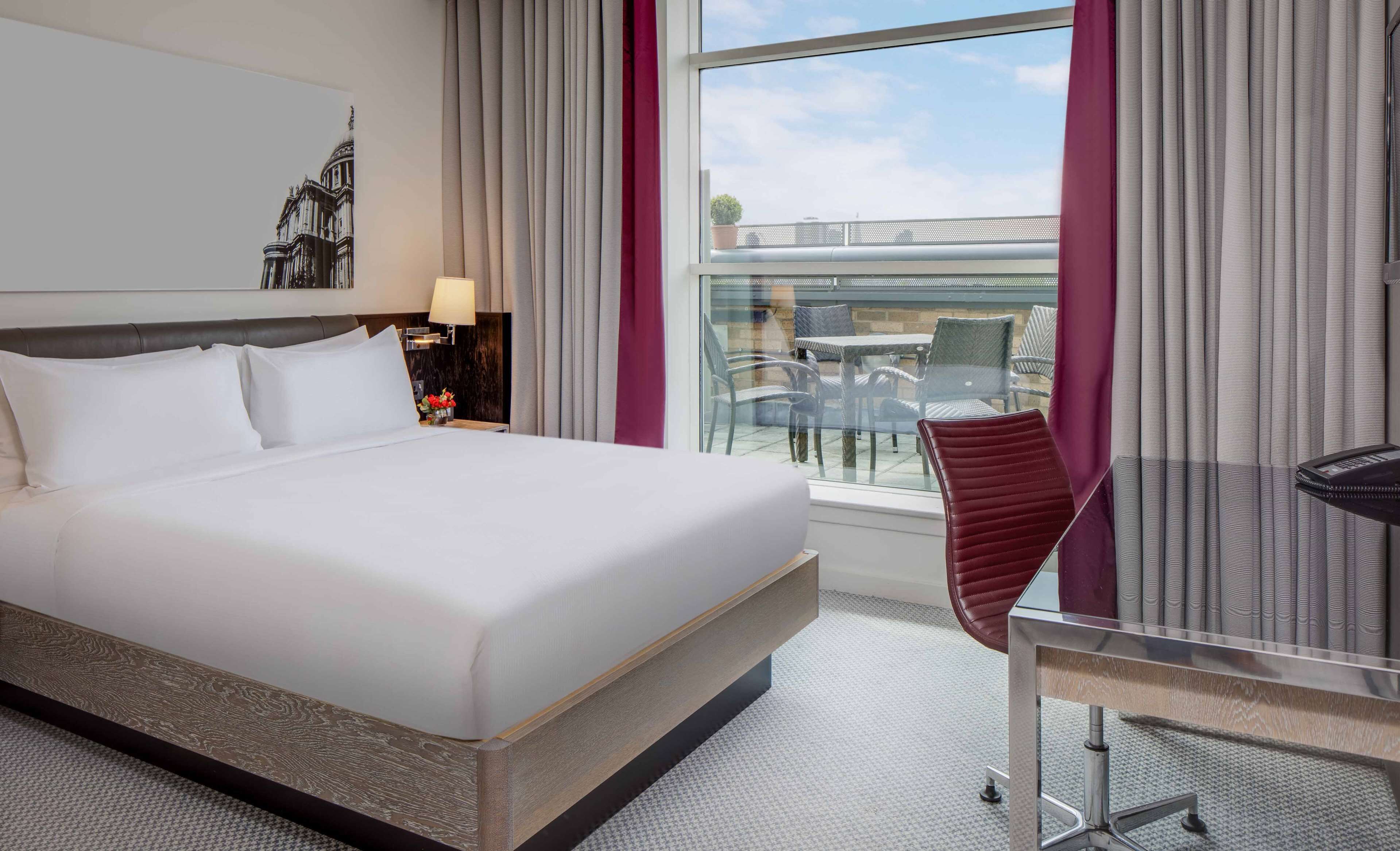 Ett hotellrum med en stor säng, ett skrivbord med en stol, ett fönster med gardiner och utsikt över utemöbler.