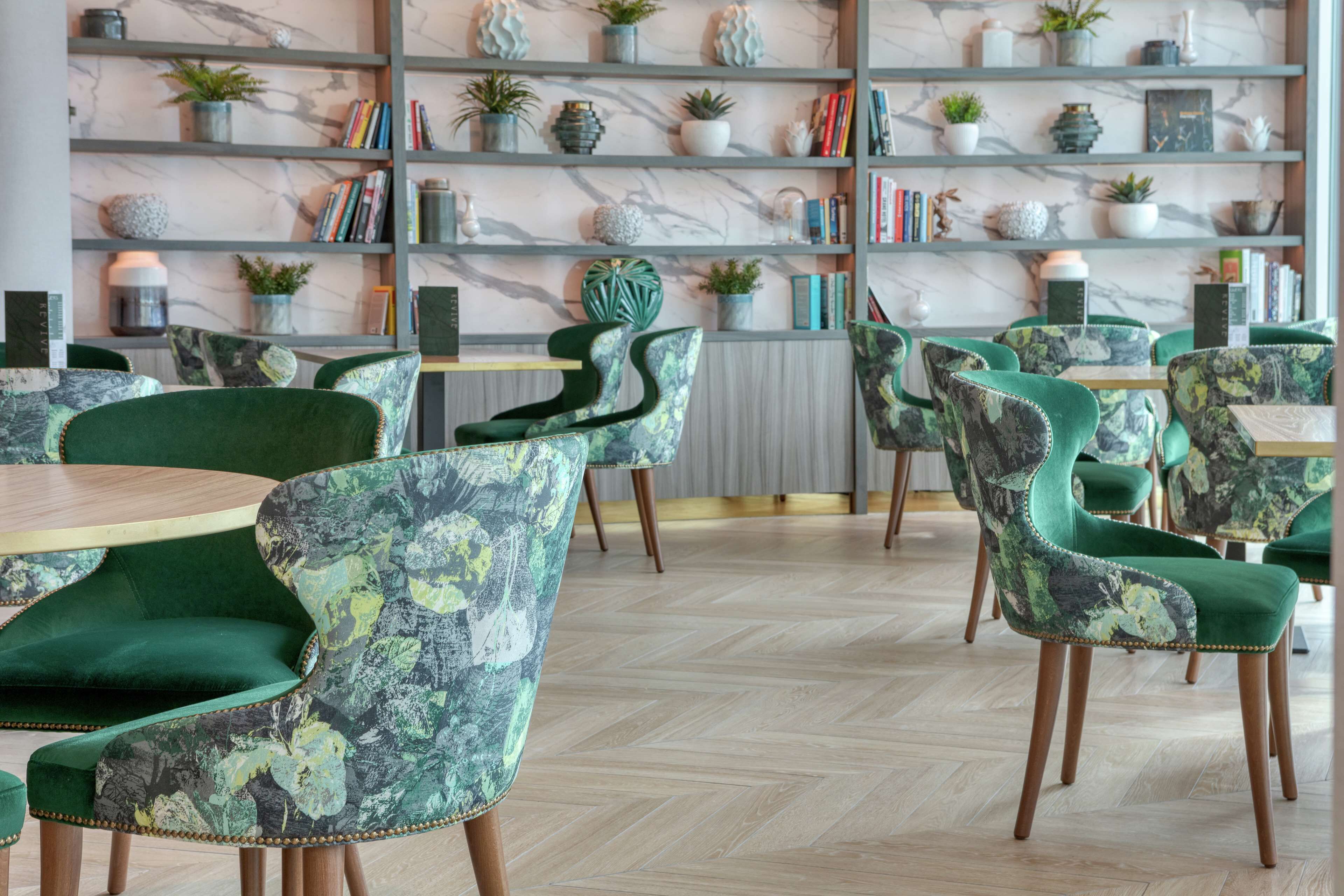 A modern interior with green upholstered chairs, marble shelves with plants and books, and wooden flooring.