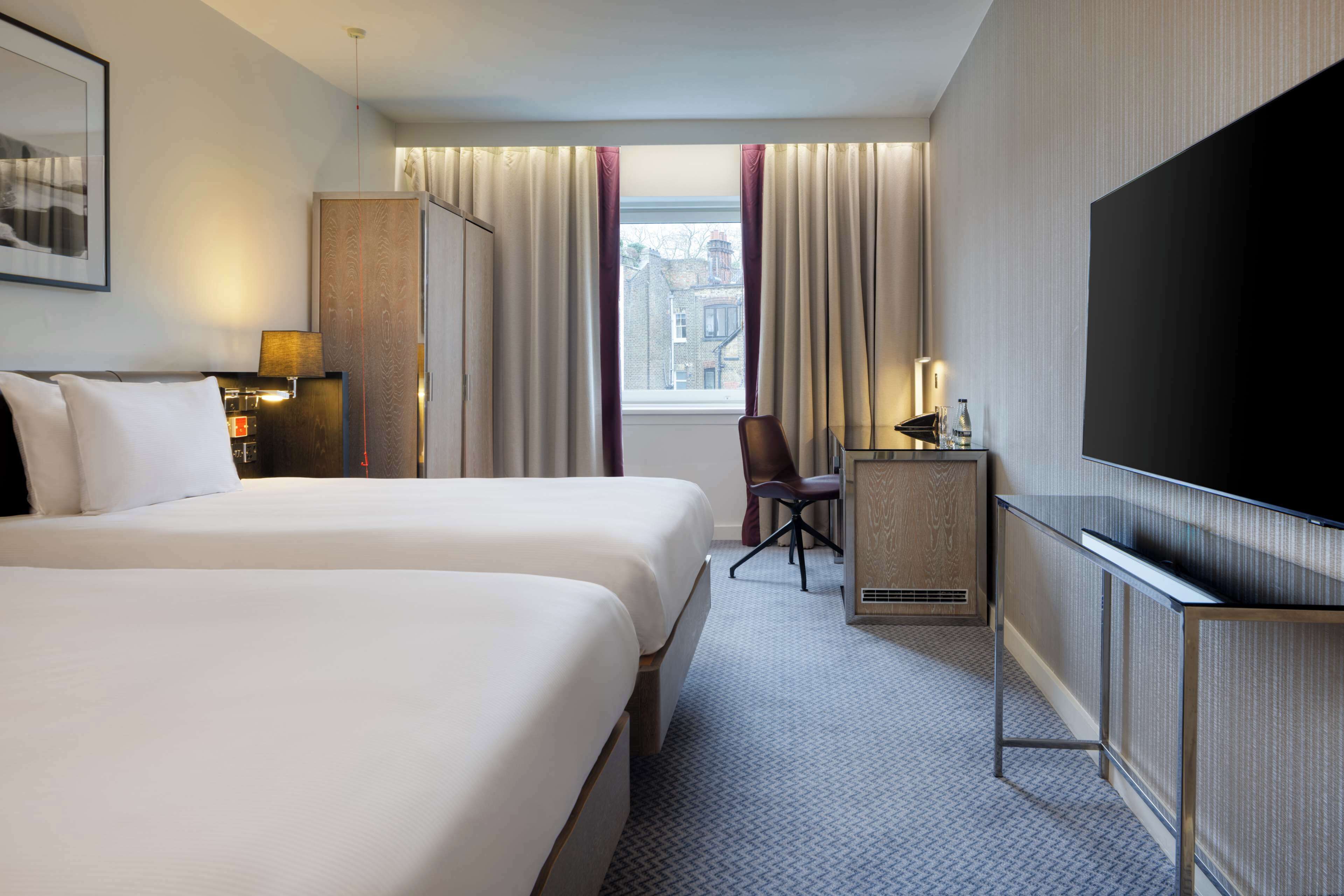 A hotel room with two beds, a desk, a TV, and a window with curtains.