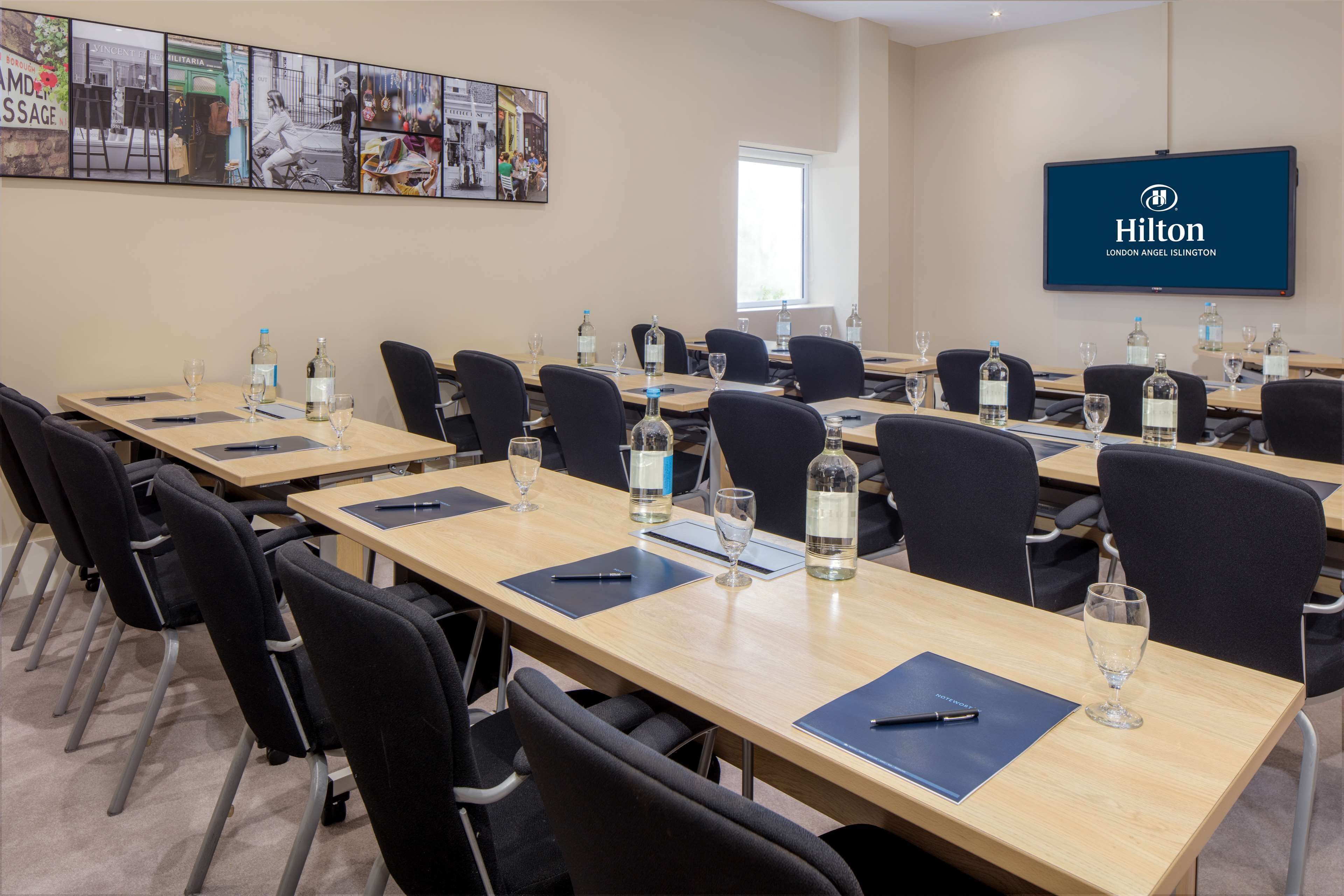 A conference room with multiple tables, chairs, and a large screen displaying the Hilton London Angel Islington logo.