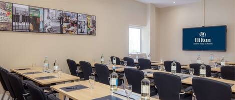 A conference room with multiple tables, chairs, and a large screen displaying the Hilton London Angel Islington logo.